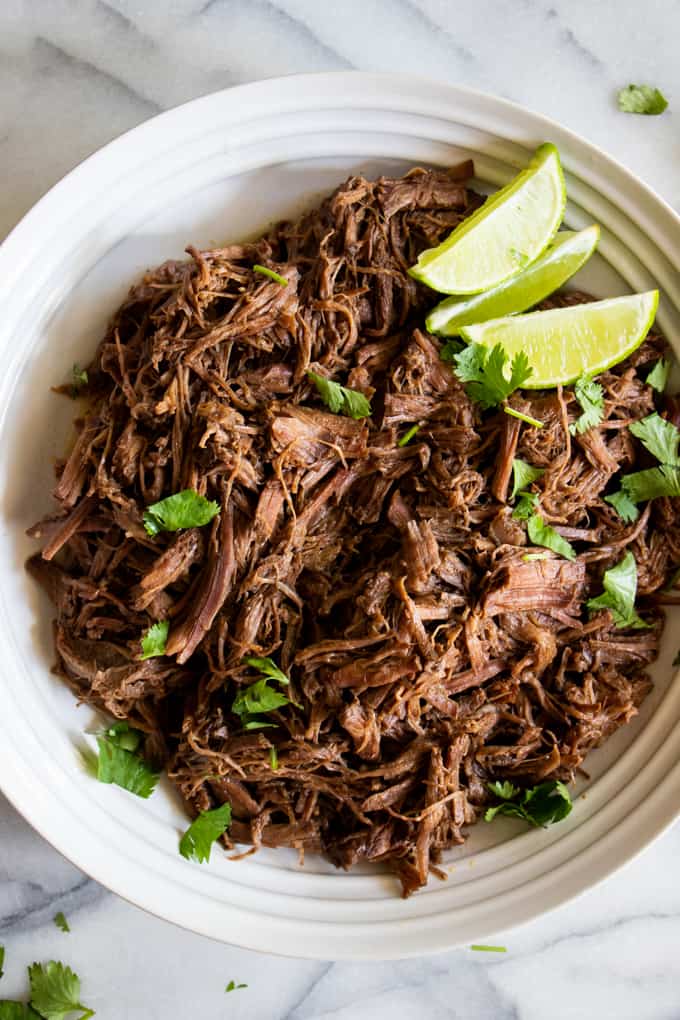 Bowl filled with shredded barbacoa topped with chopped cilantro and lime wedges.