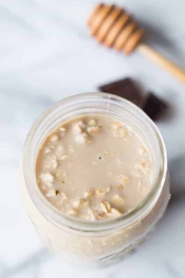 Overhead view of mocha overnight oats, with chunks of chocolate on the board in the background with a wooden honey stirrer.