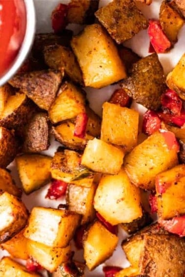 Baked, crispy breakfast potatoes mixed with red bell pepper and jalapeño spread onto a piece of parchment paper served with a cup of ketchup.