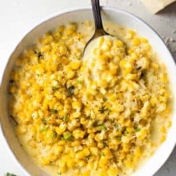 White bowl filled with cream corn, serving spoon, off to the side is a wedge of parmesan cheese.