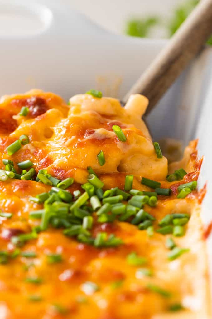 Wooden spoon digging into homemade macaroni and cheese scooping up noodles with crispy cheese topping all garnished with chopped chives.