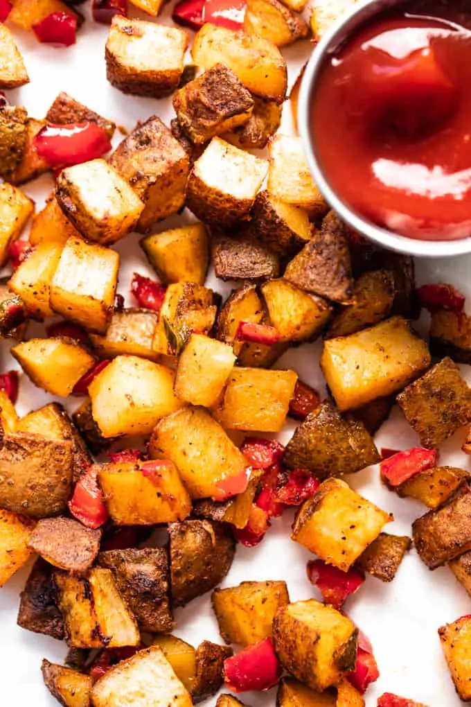 Baked, crispy breakfast potatoes mixed with red bell pepper and jalapeño spread onto a piece of parchment paper served with a cup of ketchup.