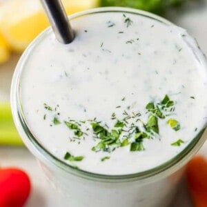 Jar filled with homemade creamy ranch dressing topped with additional chopped herbs. A spoon is dipped in the ranch and fresh veggies are on the counter around the jar.