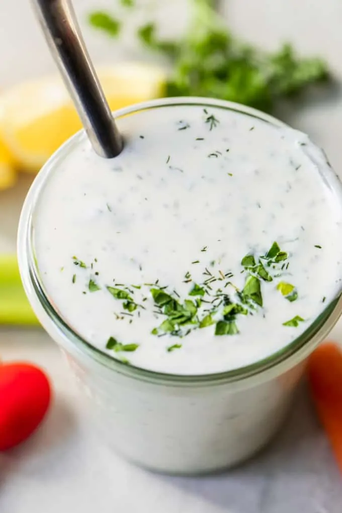 Jar filled with homemade creamy ranch dressing topped with additional chopped herbs. A spoon is dipped in the ranch and fresh veggies are on the counter around the jar. 
