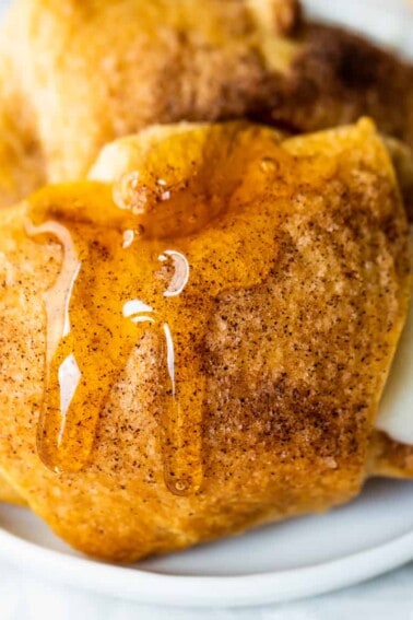 Baked sopapilla cheesecake rolls on a white plate with a drizzle of honey dripping down, a honey stirrer stick to the side.