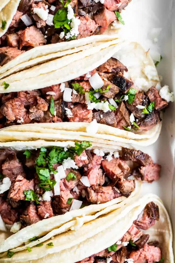 Overhead view of layered white corn tortillas that are filled with chopped grilled carne asada, topped with onion and cilantro, and crumbled queso fresco.