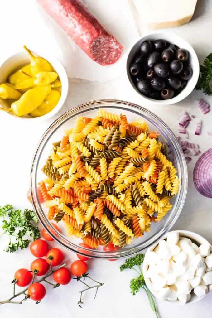 Overhead view of showing a bowl of rotini noodles, bowls of olives, mozzarella, pepperoncini, red onion, cherry tomatoes, and fresh parsley.