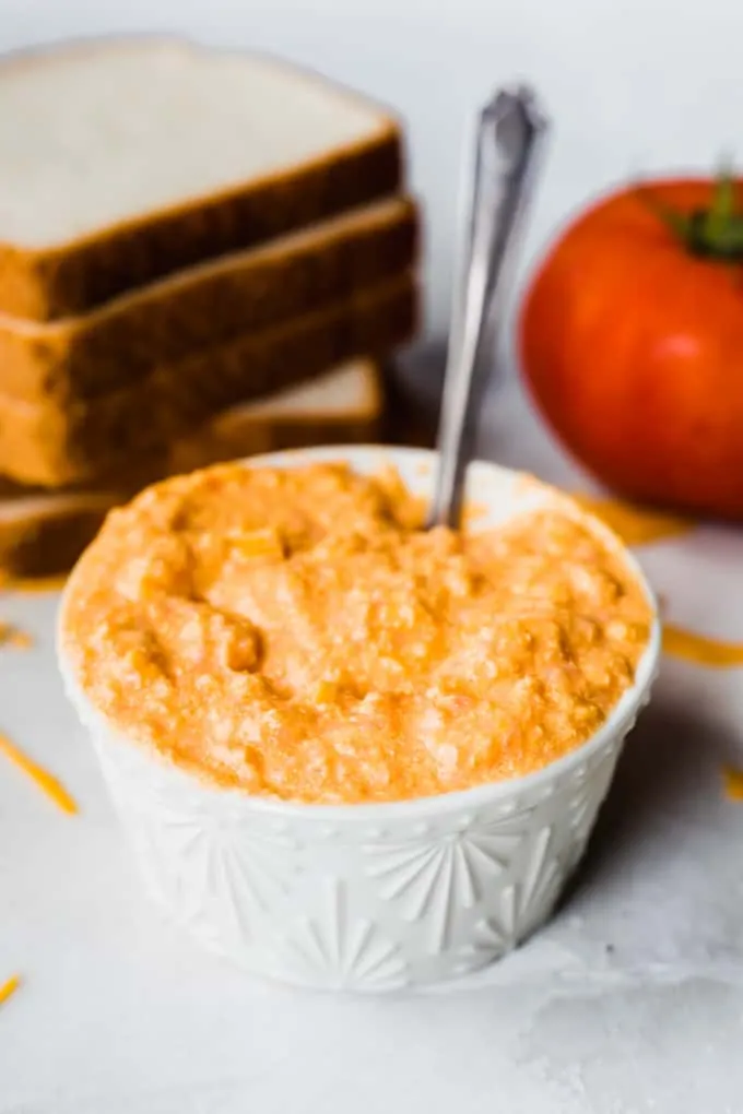Ramekin that is filled with pureed tomato with cheese and onion to make tomato sandwiches, a spoon dipped in. A stack of bread and a tomato in the background.
