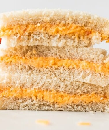 Stack of tomato twiddle sandwiches on white bread, with grated cheese on the counter around them, a tomato in the background and a ramekin filled with tomato twiddle.
