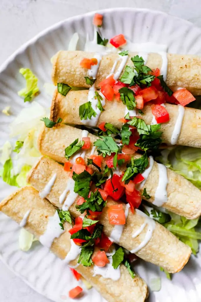 A plate filled with baked chicken taquitos, topped with drizzles of Crema, diced tomato, and fresh cilantro.