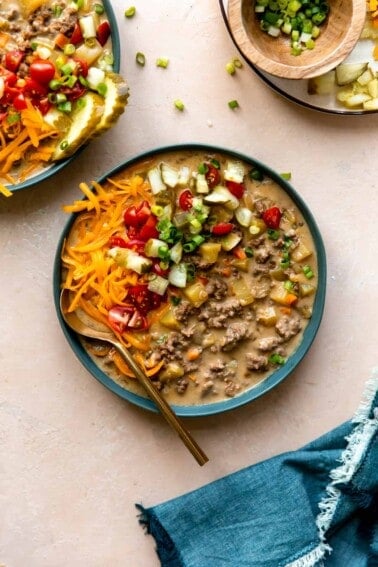 Bowl filled with cheeseburger soup and topped with diced tomatoes, pickles, cheese and herbs.