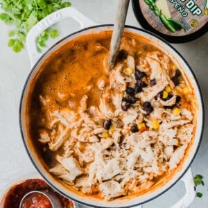 Large pot of chicken queso chili surrounded by fresh cilantro, onion, and cacique salsa and queso.