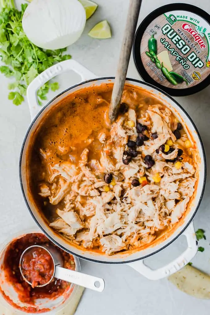 Large pot of chicken queso chili surrounded by fresh cilantro, onion, and cacique salsa and queso. 
