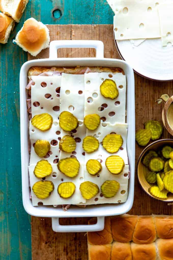 White baking dish with layers of Hawaiian rolls, sliced deli ham, sliced Swiss cheese and pickles.