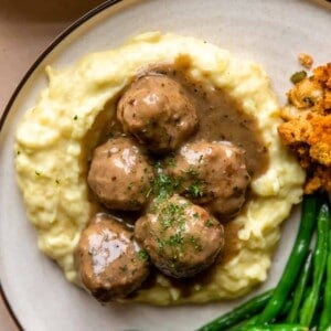 Turkey meatballs and gravy served over mashed potatoes on a plate with green beans and stuffing on the side.