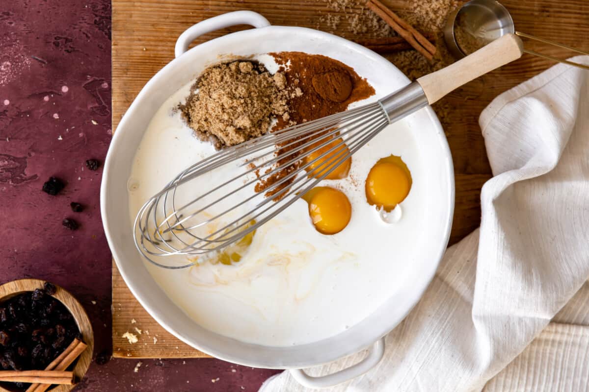 White bowl with milk, eggs, sugar, and cinnamon with a whisk.