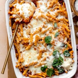 Spoon dishing into a baking dish filled with baked ziti, topped with melted cheese and basil leaves.