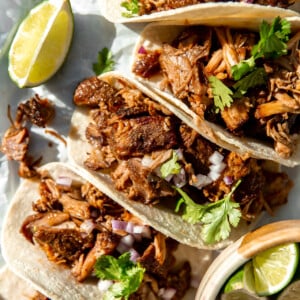 Carnitas Tacos lined up and topped with fresh cilantro, and diced red onion, lime wedges on the side.
