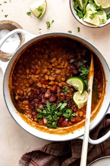 Dutch oven filled with cooked borracho beans topped with cilantro and lime wedges.