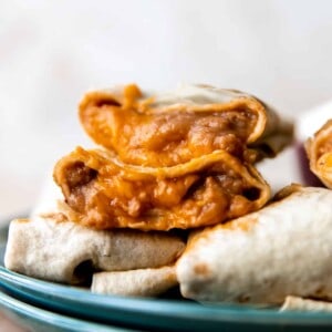 Bean and cheese burritos stacked on a plate showing melty cheese and creamy beans with red sauce inside.