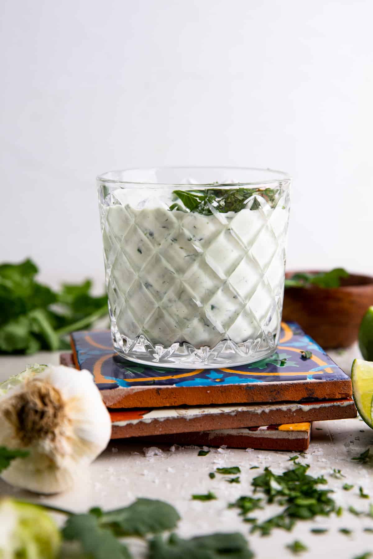 Small cup of cilantro Lime Crema on top of mexican tiles with lime wedges, garlic bulb and a bunch of cilantro in the background.
