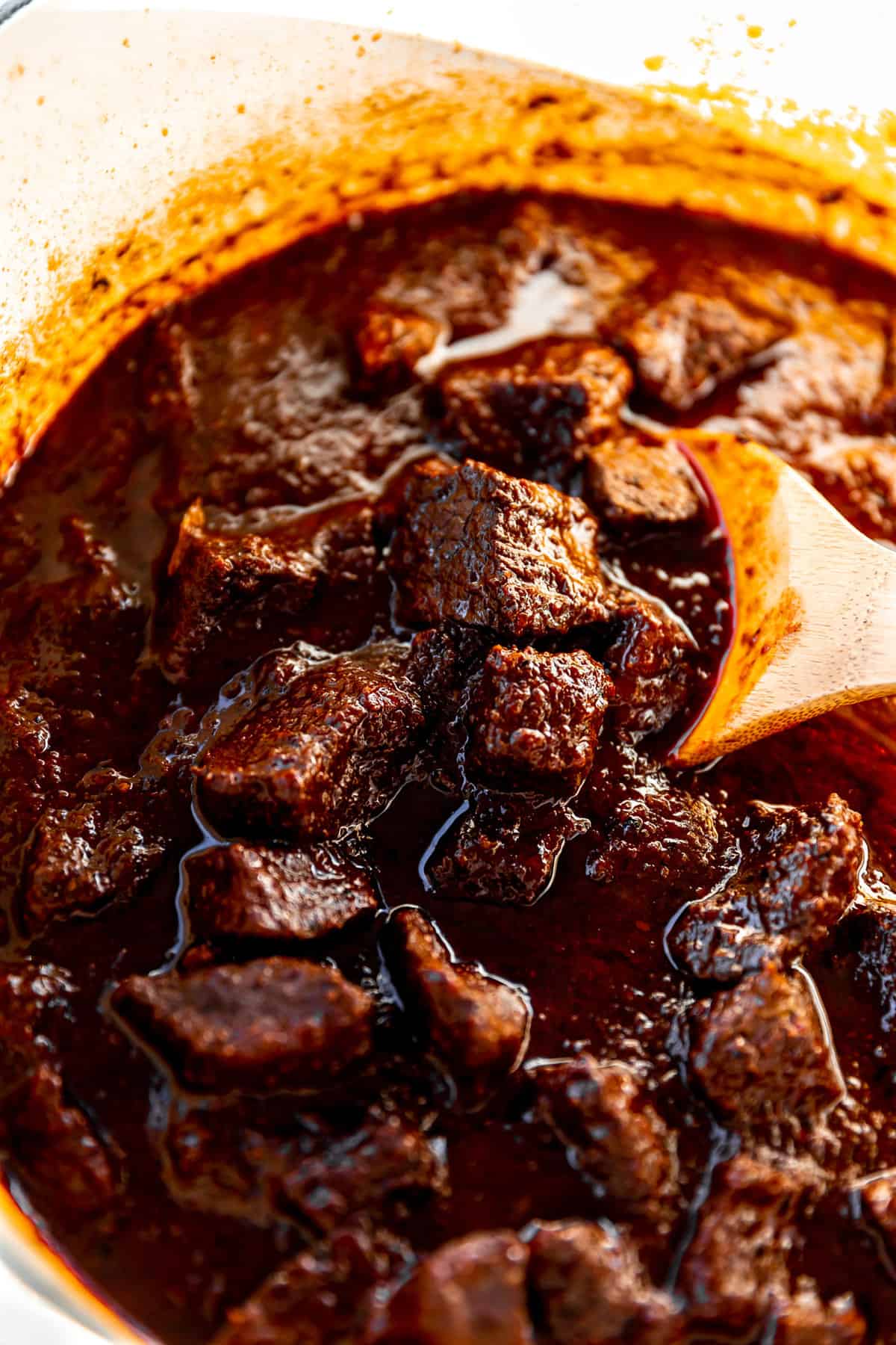 Dutch oven filled with red chile colorado being scooped up with a wooden spoon.
