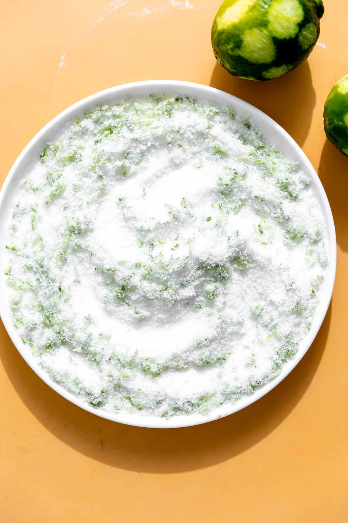 Sugar mixed with lime zest on a small plate, zested limes on the side.