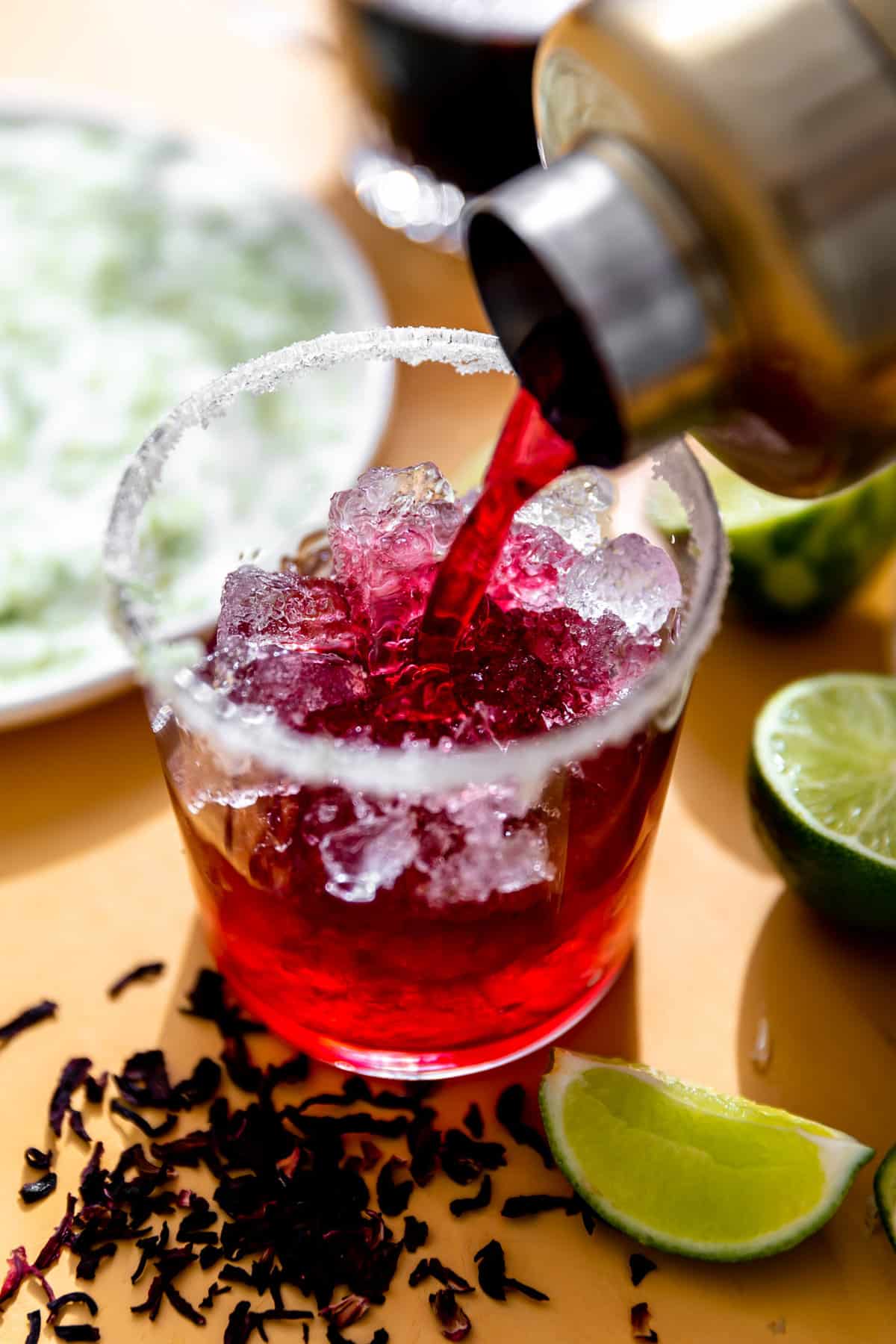 Cocktail shaker pouring out a deep pink colored cocktail into a glass filled with ice and rimmed with sugar. 