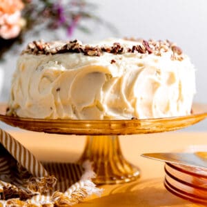 Hummingbird cake with cream cheese frosting and crushed pecans on top sitting on an orange cake stand.