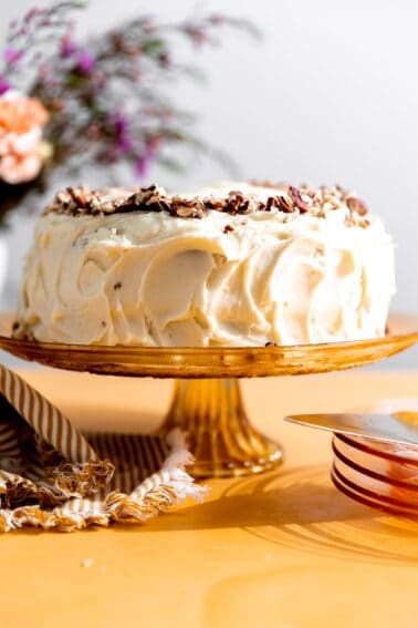 Hummingbird cake with cream cheese frosting and crushed pecans on top sitting on an orange cake stand.