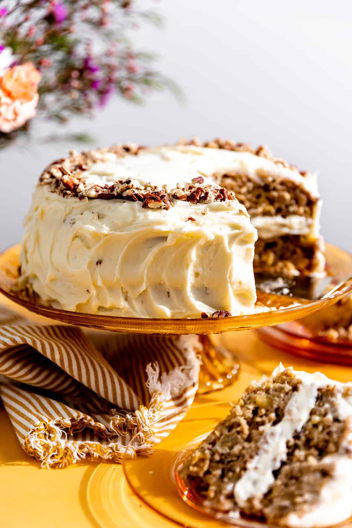 Hummingbird cake topped with pecans, on a orange glass cake stand with one slice removed and on a plate.