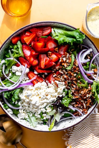 Bowl filled with salad greens topped with sliced strawberries, crumbled feta, sliced red onions and chopped pecans.