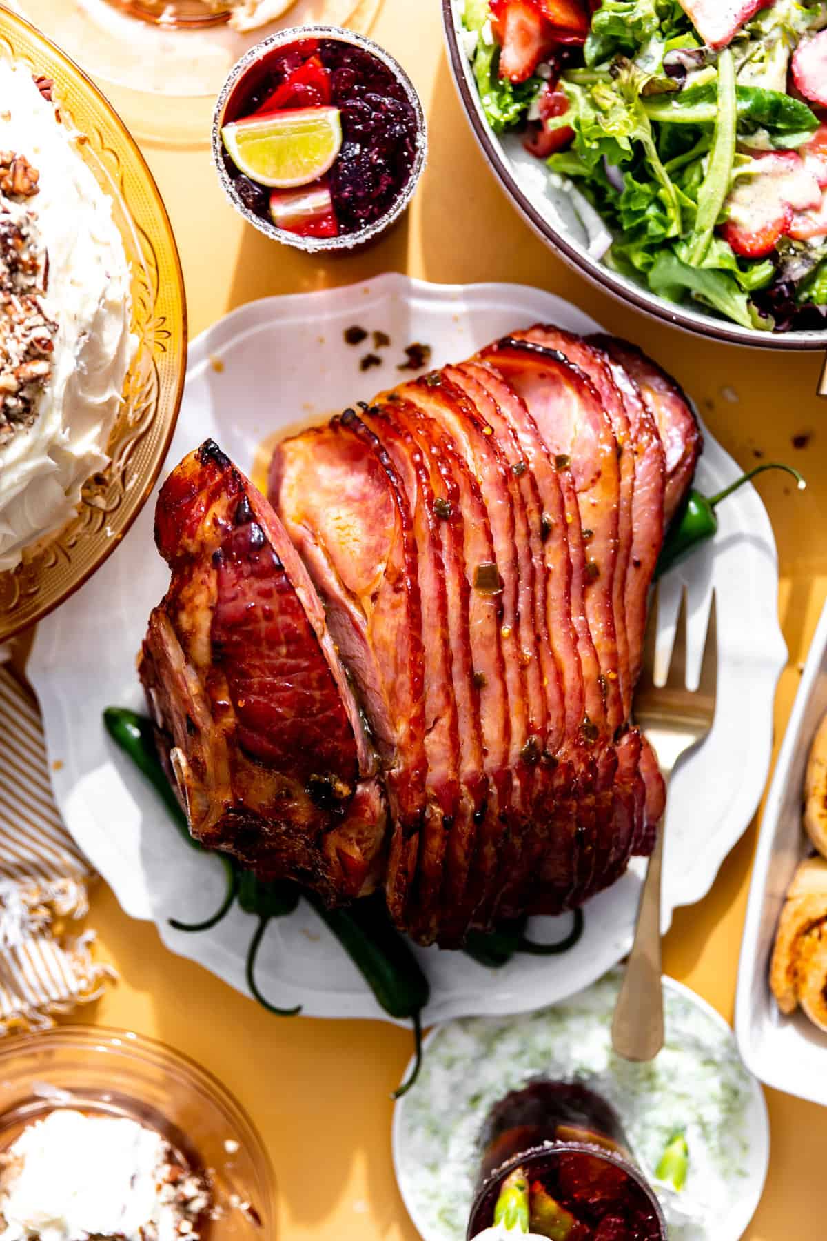 Jalapeno honey ham on a table surrounded by salad, bread rolls, margaritas and a cake. 