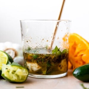 Glass filled with carnitas marinade, lime wedges, orange wedges surrounded and a gold spoon in the cup.