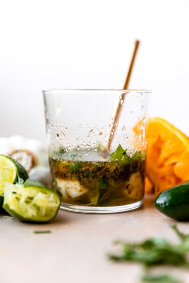 Glass filled with carnitas marinade, lime wedges, orange wedges surrounded and a gold spoon in the cup.