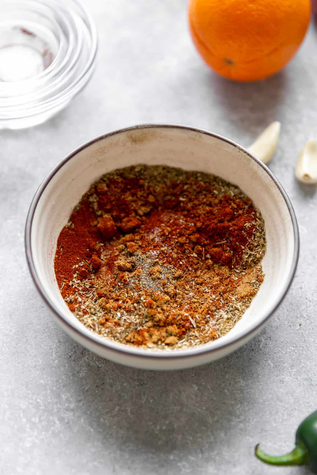 Bowl of carnitas seasoning mixed together with garlic cloves, jalapeno and an orange on the side.