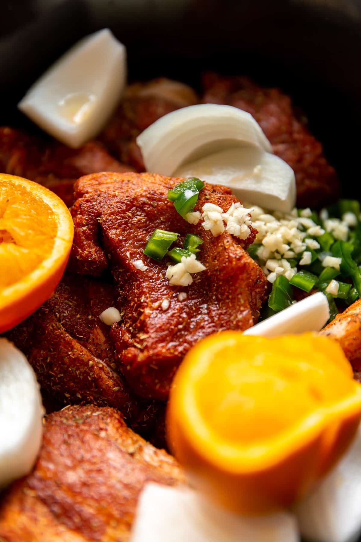 Large chunks of pork in a slow cooker with bits of onion, jalapeno, orange haves and a carnitas seasoning blend.