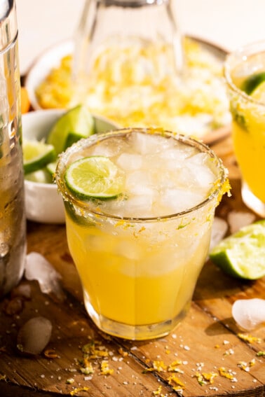 Glass filled with Texas margarita, garnished with a lime wedge, on a cutting board surrounded by sliced limes.