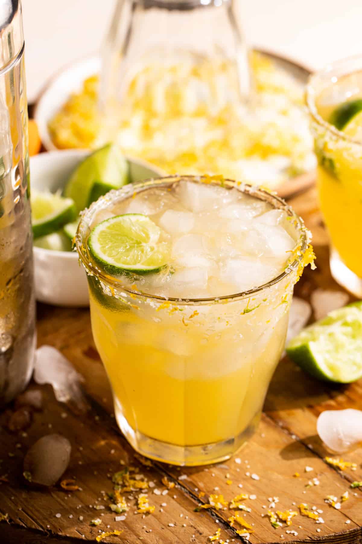 Glass filled with Texas margarita, garnished with a lime wedge, on a cutting board surrounded by sliced limes. 