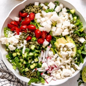 White bowl filled with chopped romaine lettuce topped with tomato, avocado, radishes, cucumber, cotija and pepitas.