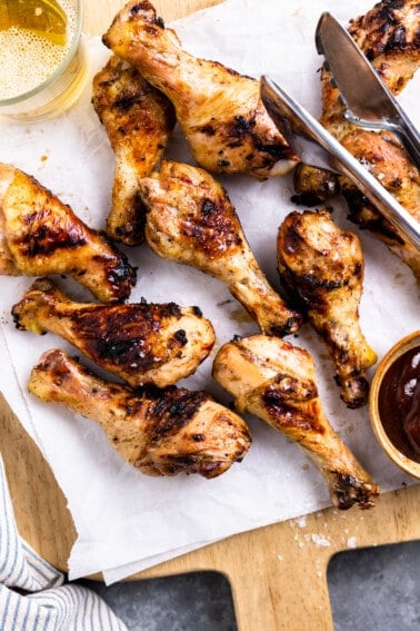 Grilled chicken legs on a wood cutting board with a bowl of bbq sauce and a beer.