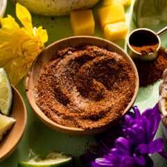 Small wooden bowl with chili lime seasoning, surrounded by chopped cucumber and fruits.