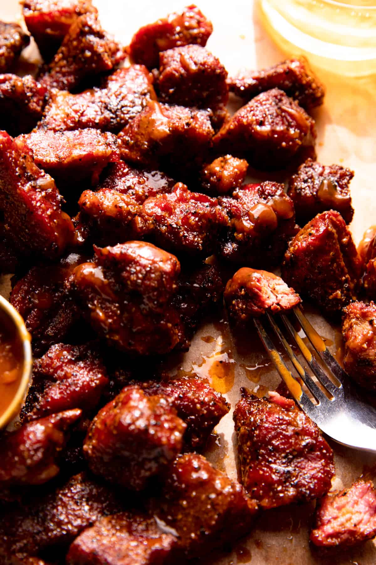 Smoked chuck roast burnt ends spread out on a piece of parchment paper with a cup of bbq sauce. 