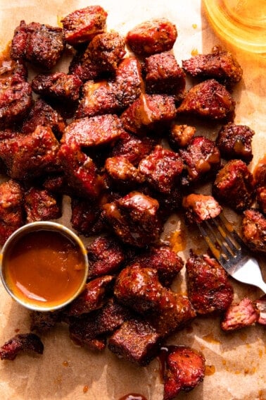 Smoked chunks of beef served with a cup of bbq sauce on a piece of brown parchment paper.