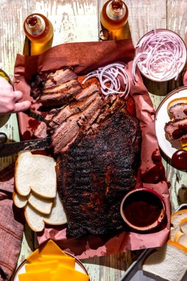 Texas BBQ spread featuring a whole brisket being sliced with white bread, cheese, red onion, and beer around it.