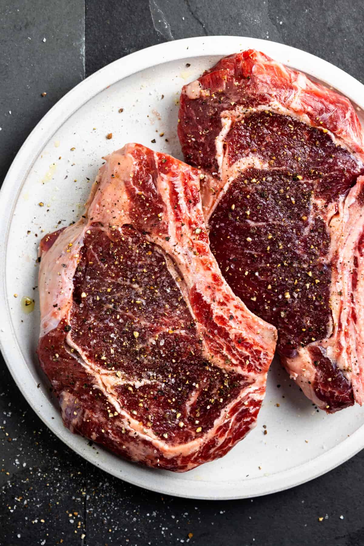 Raw ribeyes on a white plate with a seasoning of coarse black pepper and salt on them.