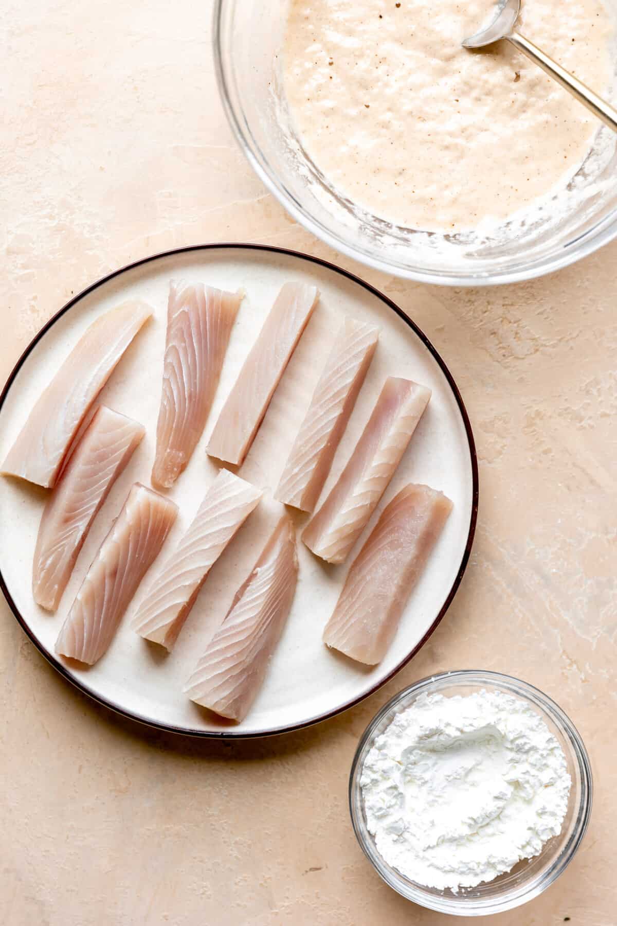 Mahi Mahi on a plate with a bowl of cornstarch on the side and beer batter in a glass bowl on the other side. 