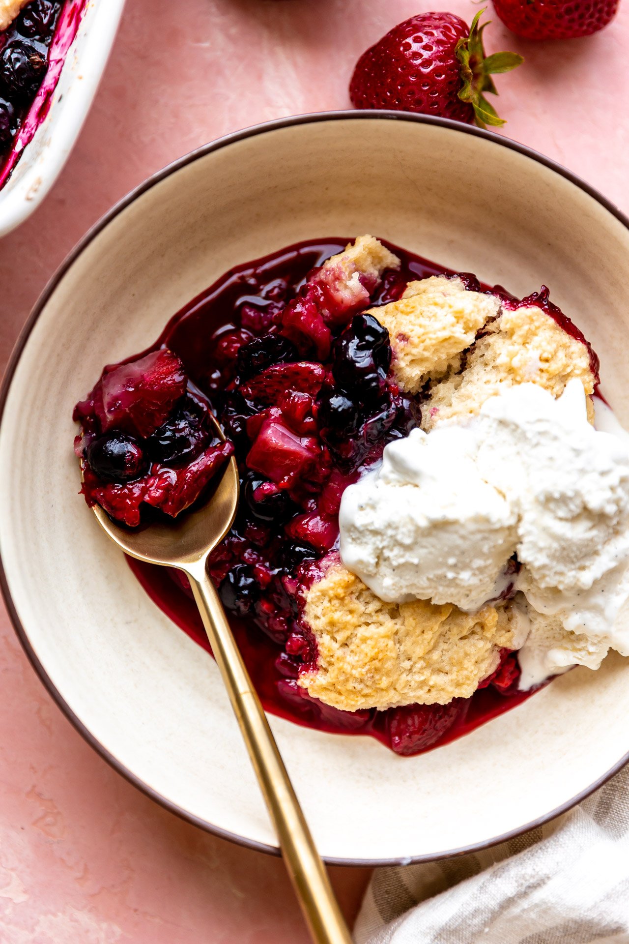 Mixed berry cobbler in a bowl topped with ice cream, being served with a spoon.
