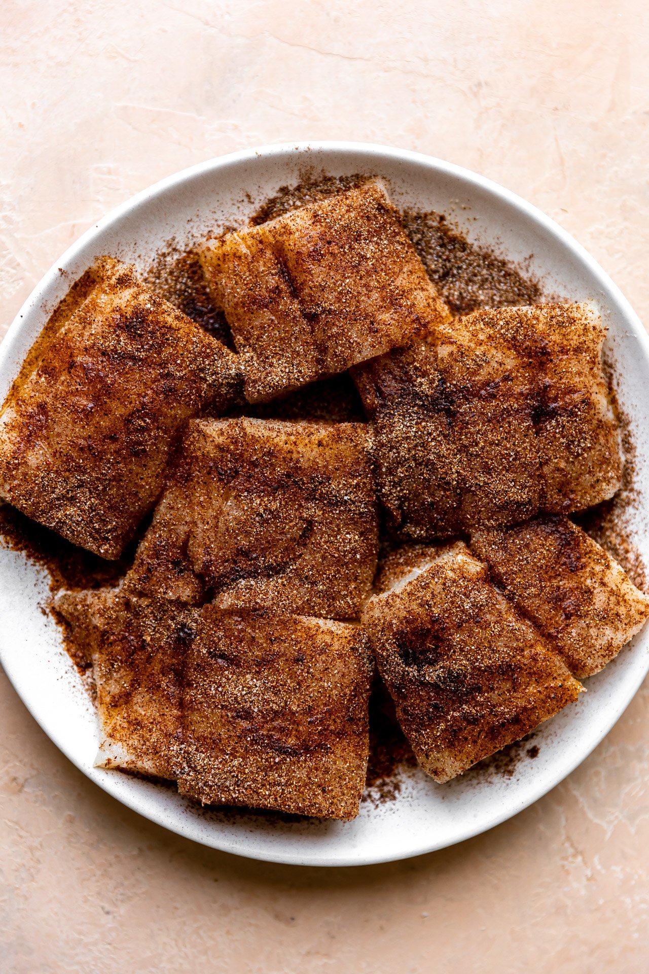 Pieces of cod on a white plate and covered with seasoning rub.