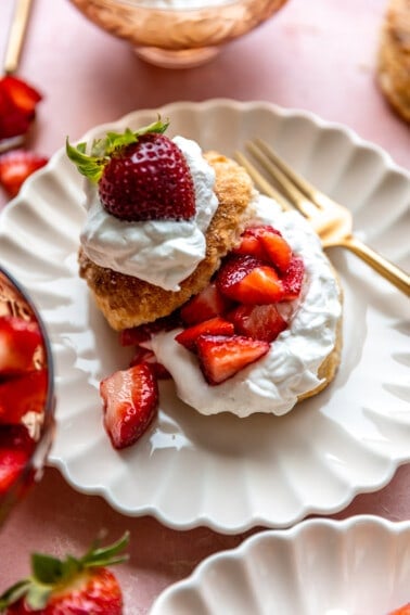 Strawberry shortcake served on a white scalloped plate, gold fork on the side.